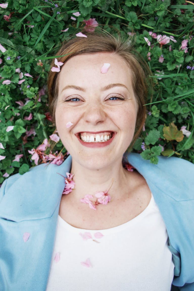 A cheerful woman lies in grass with flowers, enjoying a peaceful outdoor moment.