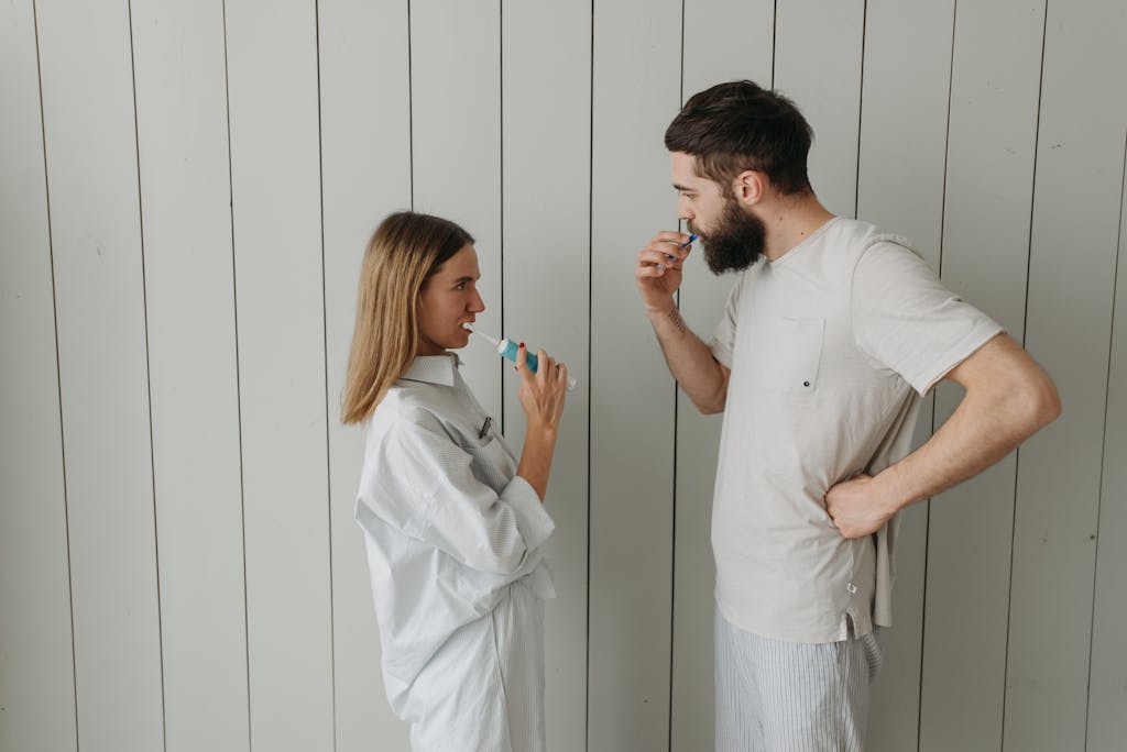 A couple brushing their teeth indoors, sharing a morning routine. Side view highlights interaction.