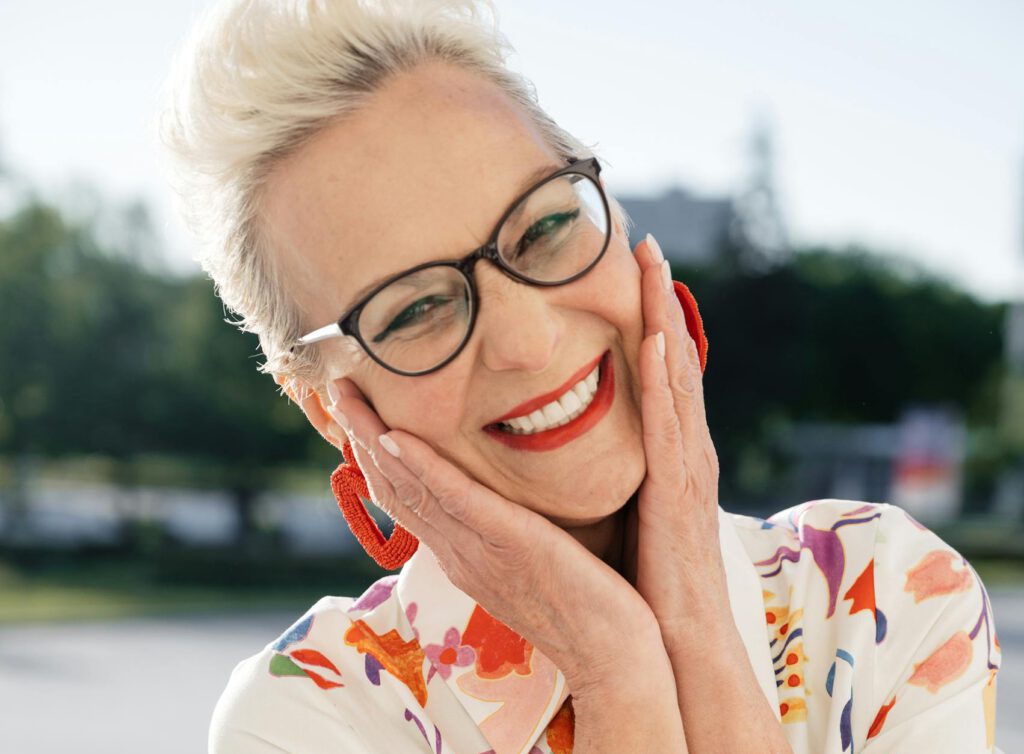 Elderly woman with eyeglasses smiling and posing outdoors in a floral dress.